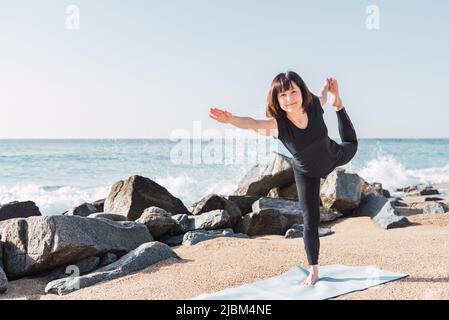 Frau in Sportkleidung balanciert auf dem Bein in Natarajasana auf der Matte. Beim Yoga an der sandigen Küste in der Nähe des Meeres an sonnigen Tag und Blick auf die Kamera Stockfoto