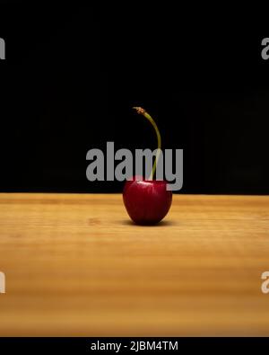 Single RIPE Cherry platziert auf einem Holzschneidebrett auf einem dunkelschwarzen Hintergrund. Stockfoto