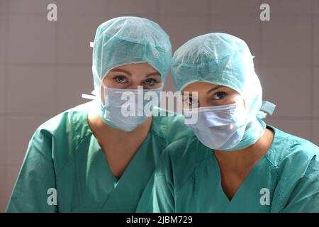 Zwei junge Frauen werden in einem Operationssaal mit medizinischer OP-Kleidung gesehen. Beide tragen geschützte Gesichtsmasken über Mund und Nase. Stockfoto