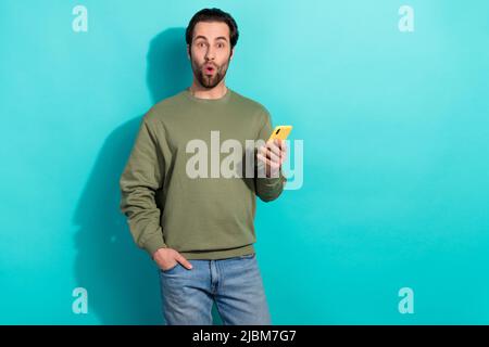 Foto von beeindruckt Brunet jungen Kerl halten Telefon tragen khaki Sweatshirt isoliert auf blauem Hintergrund Stockfoto