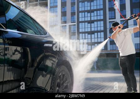 Mann mit Schutzkappe, der sein schwarzes modernes Auto mit Hochdruckwasser in Betrieb genommen hat. Waschen von Auto mit fließendem Wasser auf dem Hintergrund des Wohngebäudes. Stockfoto