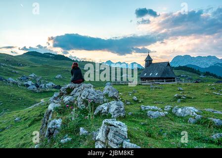 Einsamer Reisender sitzt auf Felsen, fasziniert von Grasland Berge, Hütten, Holzkirche die "Kapelle unserer Lieben Frau vom Schnee" velika planina Stockfoto