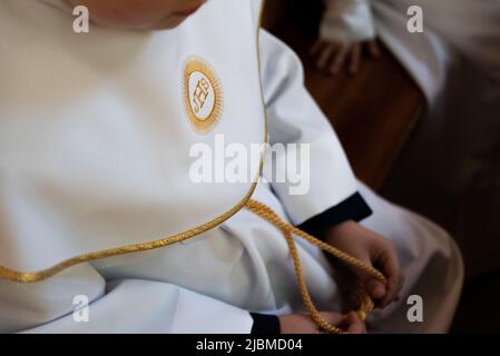 IHS christliches Symbol auf einem Kommunionskind. IHS Christogramm bestickt mit Goldfaden auf einer weißen Alba. Kleid der ersten Heiligen Kommunion. Stockfoto