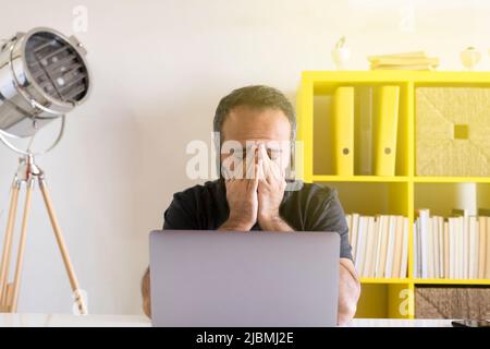 Gestresster junger Mann, Vorderansicht des gestressten jungen Mannes, Gesicht mit Händen bedeckend. Im Büro, mit Laptop arbeiten, müde oder traurig oder deprimiert. Stockfoto