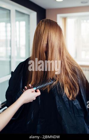 Weibliche Kundin mit langen, geraden, schönen Haaren lacht, während ein Friseur vor ihrem Gesicht Stränge herauskämmt und im Schönheitssalon arbeitet Stockfoto