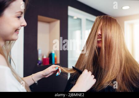 Weibliche Kundin mit langen, geraden, schönen Haaren lacht, während ein Friseur vor ihrem Gesicht Stränge herauskämmt und im Schönheitssalon arbeitet Stockfoto