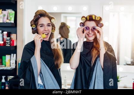 Zwei lustige junge Freundinnen in Lockenwicklern tragen Umhänge und haben Spaß zusammen im Schönheitssalon. Freundinnen, die mit Rollen herumtollten, machten Stockfoto