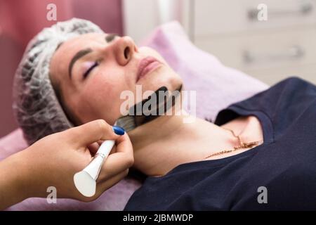 Auftragen einer Creme-Maske für das Carbon-Peeling auf das Gesicht einer erwachsenen Frau. Stockfoto