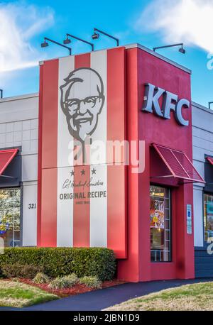 Die Außenfassade des KFC Fast Food-Restaurants mit frittierten Hühnchen und das Logo an einem hellen, sonnigen Tag mit Sträuchern, blauem Himmel und Außenbeleuchtung. Stockfoto