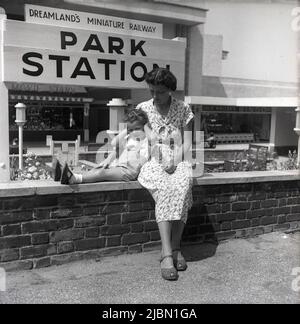 1950s, historisch, eine Mutter und ihr junger Sohn sitzen auf einer kleinen Wand an der Park Station in Dreamlands Miniature Railway, im Dreamland Pleasure Park, Margate, Kent, England, Großbritannien. Stockfoto