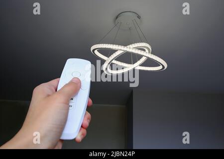 Moderner LED-Kronleuchter an der Decke. Die Frau schaltet das Licht mit der Fernbedienung ein. Stockfoto