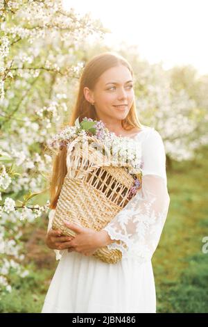 Junge attraktive Frau von natürlicher Schönheit mit Blumenkorb im Sommergarten, die die blühende Natur des Frühlings genießt Stockfoto