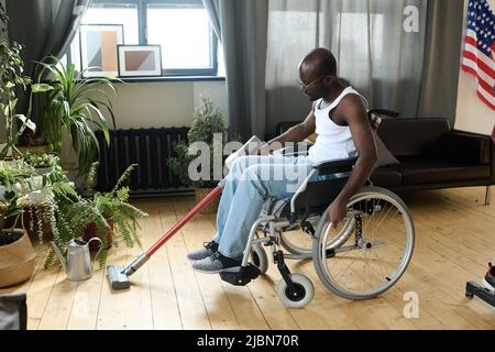 Afroamerikanischer junger Mann mit Behinderung, der im Rollstuhl sitzt und Staubsauger zur Hausarbeit verwendet Stockfoto
