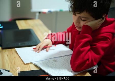 Bezaubernder, vorpubertär-kaukasischer Schuljunge, der am Holzschreibtisch sitzt und sich auf Hausaufgaben konzentriert. Bildung, Schulleben, Gelehrsamkeit, Wissen Co Stockfoto