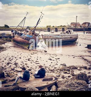 Zwei Jungen essen Fisch und Chips vor zwei zerstörten Fischtrawlern, Fleetwood, Großbritannien Stockfoto