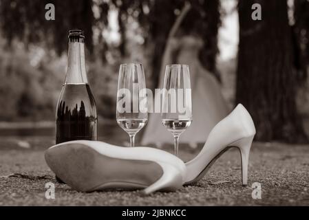 Eine Champagnerflasche, ein Paar High Heels und zwei Sektgläser befinden sich auf dem Boden im Freien, Ende einer Hochzeit, Schwarz-Weiß-Foto Stockfoto