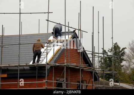 Zwei Männer, die zum Schutz des Dachs mit einer Leiter und weggeworfenen Isolationsmaterialien auf dem Dach des Neubaus Kacheln verlegen Stockfoto