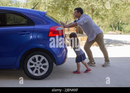 Bild eines Mannes, der mit seiner Tochter ihr Auto schiebt, nachdem er eine Panne hatte und das Benzin ausging. Verweis auf den aktuell teuren Kraftstoff Stockfoto