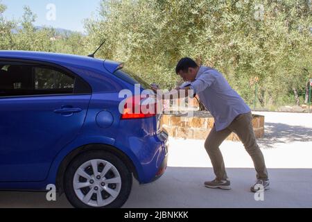 Bild eines Mannes, der sein Auto schiebt, nachdem es zusammengebrochen war und das Benzin ausgegangen war. Verweis auf den aktuell teuren Kraftstoff Stockfoto