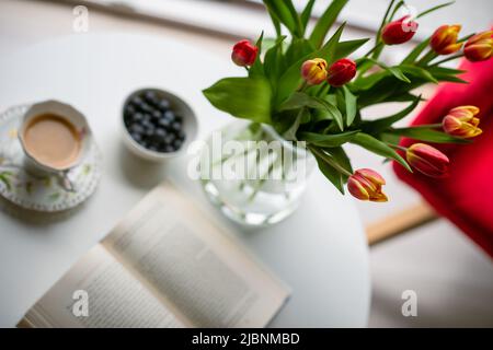 Frische Frühlingsblumen in Glasvase und eine Tasse Kaffee mit einem Buch zum Lesen Stockfoto