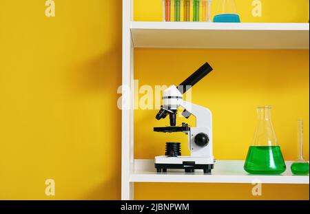 Regaleinheit mit verschiedenen Laborglas- und Mikroskopen in der Nähe der gelben Wand Stockfoto