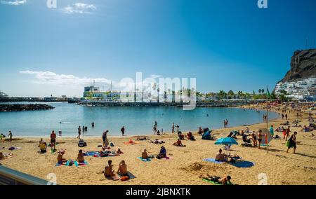 Februar 02 2022 Playa de Mogan auf gran canaria ein sehr schöner Ort, der von vielen Touristen besucht wird, die am Strand auf dem gelben Sand und im Bac sitzen Stockfoto