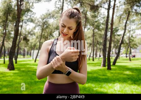 Junge, schöne Frau, die im Stadtpark sportliche Kleidung trägt und ihr schmerzhaftes Handgelenk im Freien hält. Leiden Schmerzen an Händen und Fingern, Arthritis Inflam Stockfoto