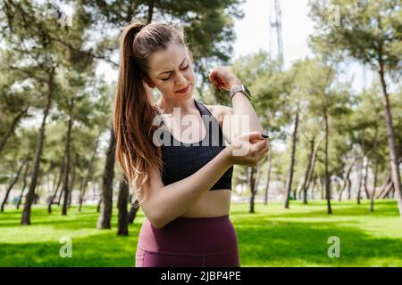 Porträt einer jungen Brünette Frau, die einen Sport-BH im Stadtpark trägt und an chronischem Gelenkrheumatismus leidet. Ellenbogenschmerzen und Behandlung Stockfoto