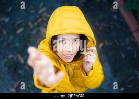 Frau, die die Kamera anschaut, trägt einen gelben Mantel mit gelber Kapuze, wobei die Hand nach der Kamera greift. Stockfoto