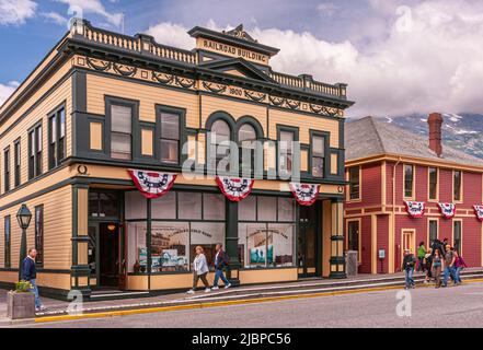 Skagway, Alaska, USA - 20. Juli 2011: Gelb-grünes Gebäude der hölzernen Eisenbahn auf der 2. Ave unter blauer Wolkenlandschaft. Drapierte Flaggen, Sichtfenster mit poste Stockfoto