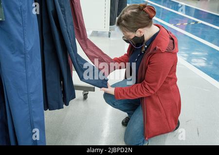 Frau in Maske gegen Virus wählt dunkelblaue und rote Hosen im Bekleidungsgeschäft Stockfoto