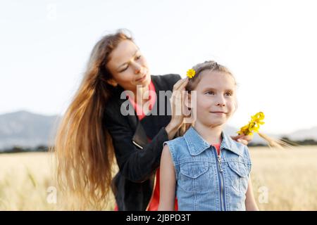 Mutter flechtet die Haare der Tochter mit gelben Blüten auf dem Feld. Stockfoto