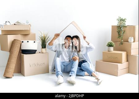 Multirassische stilvolle Paar in der Liebe, junge Familie, sitzen sie an einer weißen Wand, zwischen Kartons, in ihrem neuen Haus, zeigt das Dach mit ihren Händen, Blick auf die Kamera, lächelnd Stockfoto