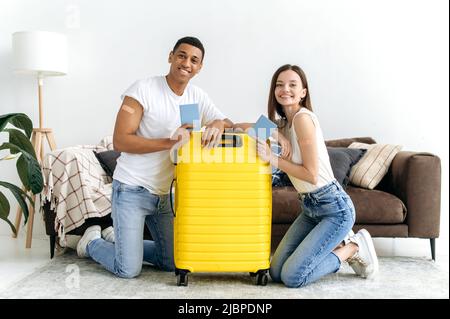 Geimpfte Typen und Mädchen verschiedener Nationalitäten, mit ihren Pässen, sitzen auf dem Boden im Wohnzimmer neben dem Koffer mit Dingen, gehen nach der Pandemie in den Urlaub, lächeln glücklich Stockfoto