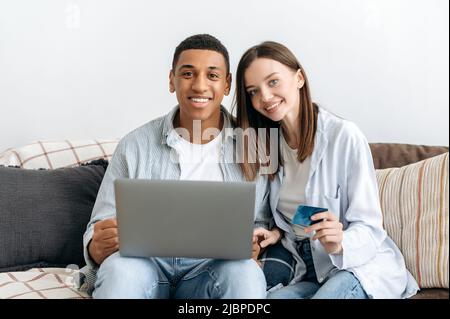 Online-Shopping. Glückliches, positives gemischtes Familienpaar, das auf einem Sofa im gemütlichen Wohnzimmer sitzt, Laptop und Kreditkarte zum Online-Shopping nutzt, Dinge für zu Hause kauft, Lieferung nach Hause bestellt, lächelt Stockfoto
