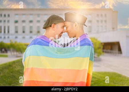 Ein Paar von verliebten schwulen Männern umarmt sich gegenseitig, eingewickelt in eine schwule Flagge, in einem Park bei Sonnenuntergang, mit Reflexen der Sonne. Konzept von Vielfalt, Stolz, Stockfoto