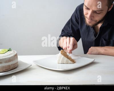 Ein Mann isst ein Stück vegane Zitronenkuchen mit Mandel, Kokosnuss und Zitronenbasis mit Zitronencreme-Buttercreme auf einem weißen Tisch und einem weißen Rücken Stockfoto