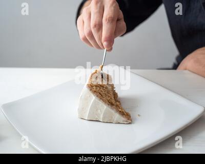 Eine Männerhand schneidet ein Stück vegane Zitronenkuchen mit Mandel, Kokosnuss und Zitronenbasis mit Zitronencreme-Buttercreme auf einem weißen Tisch und einem Whi Stockfoto