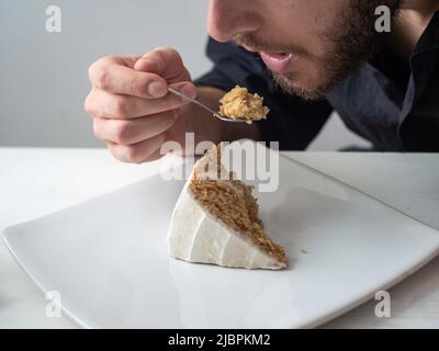 Ein Mann isst ein Stück vegane Zitronenkuchen mit Mandel, Kokosnuss und Zitronenbasis mit Zitronencreme-Buttercreme auf einem weißen Tisch und einem weißen Rücken Stockfoto