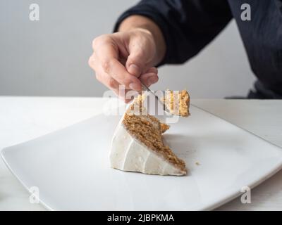 A man's Hand schneidet und hält ein Stück vegane Zitronenkuchen mit Mandel, Kokosnuss und Zitronenbasis mit Zitronencreme-Buttercreme auf einem weißen Tisch Stockfoto