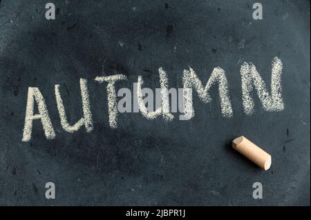 HERBST. Der Name der Saison ist in gelber Kreide auf einer schwarzen Tafel geschrieben. Handgeschriebener Text. Daneben liegt ein Stück farbige Kreide. Stockfoto