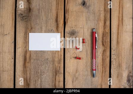 Leere Visitenkarte, Stift und Stecknadeln vor einem hölzernen Hintergrund. Ein weißes Papierrechteck, drei Stecknadeln und ein Kugelschreiber liegen auf dem alten c Stockfoto