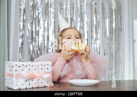 Das charmante, festlich gekleidete, rothaarige Baby, das 4-5 Jahre alt ist, genießt zu ihrem Geburtstag einen leckeren Kuchen und sitzt am Tisch im Café neben der Geschenkbox. Kinder Stockfoto