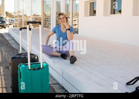 Blonde lächelnde und attraktive Frau, die mit Gepäck auf dem Boden des Busses oder Bahnhofs sitzt. Warten auf Transport Stockfoto