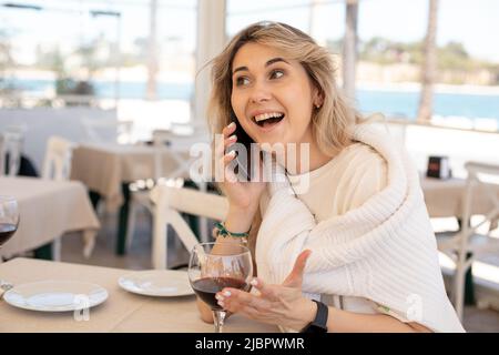 Die junge, überraschte Frau trägt ein weißes T-Shirt und sitzt am Tisch in der Nähe eines Weinglases im Restaurant und spricht auf dem Smartphone. Stockfoto
