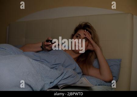 Porträt einer jungen müden erschöpften Frau, die auf einem beigefarbenen Bett liegt und den Kopf an der Hand lehnt, die Fernbedienung hält und fernsieht. Stockfoto