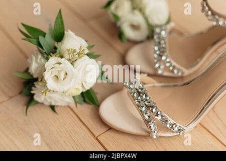 Elegante Schuhe für Hochzeitsfrauen mit Absätzen und einem Strauß frischer weißer Rosen für die Braut auf dem Boden im Zimmer. Weibliche Accessoires Nahaufnahme, beige Hintergrund Stockfoto