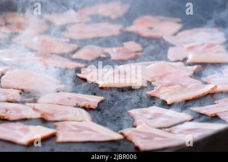 Nahaufnahmen von einer großen Menge Speck, der auf einer schwarzen grillroste oder einem heißen Teller kocht und brutzelt. Rauch verwischt den Hintergrund teilweise als Bac Stockfoto
