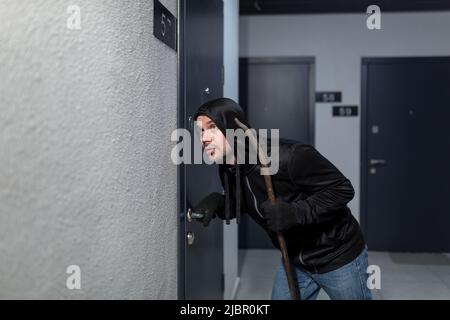 Der Räuber versucht, das Türschloss in der Wohnung zu brechen, in Schwarz gekleidet Stockfoto