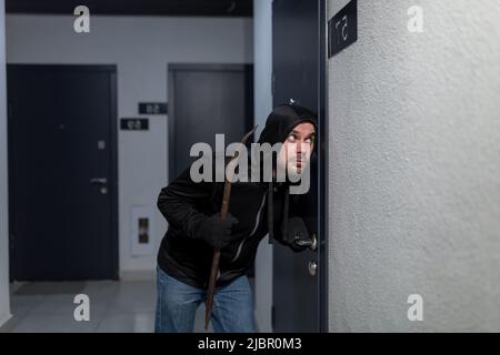 Der Räuber versucht, das Türschloss in der Wohnung zu brechen, in Schwarz gekleidet Stockfoto